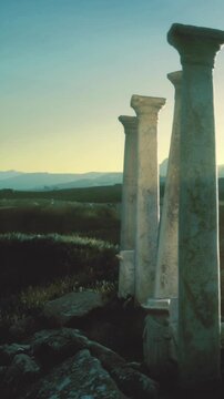 Four weathered columns rise in the landscape, surrounded by grass. The sun sets in the background, casting a warm light on the ruins. This location shows signs of history and time.