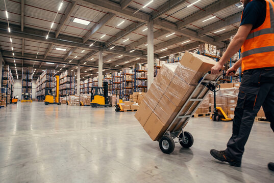 Warehouse worker pushing hand truck loaded with cardboard boxes in large distribution center