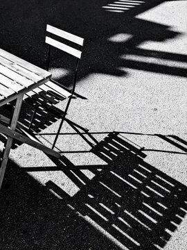 High contrast black and white abstract composition of a folded wooden chair and its strong, striped diagonal shadow on urban asphalt.