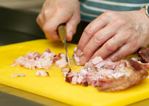 Hands of a person chopping diced meat on a yellow cutting board with a knife, showcasing preparation for cooking in a kitchen environment with stainless steel surfaces visible