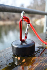 Fototapeta premium A red carabiner secures a strong magnet with a rope, resting on a wet wooden surface near water.