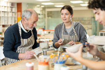 Fototapeta premium Young woman, young man and elderly man in ceramic workshop sculpting clay products during master class