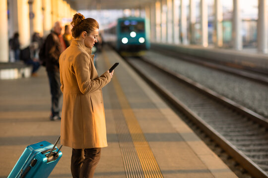 A woman stands on a station platform with her suitcase, checking her phone as a train arrives on the tracks in the background.