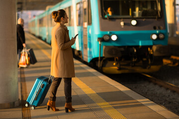 Standing on a sunny railway platform, a woman in a tan coat manages her travel plans on a...