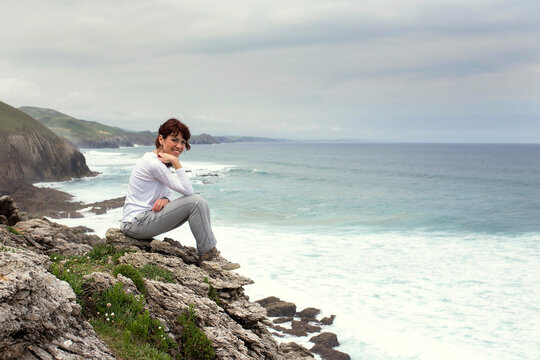 Side view of a smiling woman in casual clothing sitting on rocks by the sea, Santillana del Mar, Cantabria, Spain