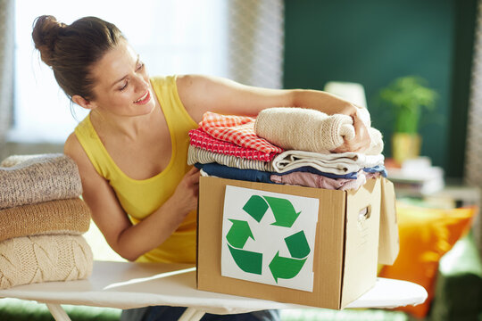 A smiling woman in a yellow top places folded knitwear into a recycling box on an ironing board, exemplifying sustainable wardrobe management and decluttering.