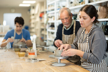 Mature man teacher shows girl how to sculpt ceramic product from raw clay in workshop