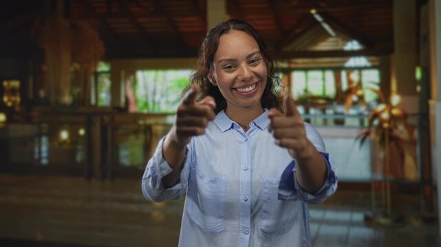 Woman points finger at camera with hands visible and a bright smile inside a hotel building lobby; confidence welcome.