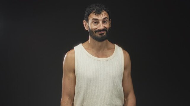 Man with beard in white tank top tilts head and shrugs shoulders, showing bare arms and a sly smirk under studio sidelight; bemusement.