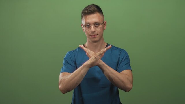 Man doctor in blue scrubs makes arms crossed x gesture with hands and forearms visible against studio green backdrop; denial protest.