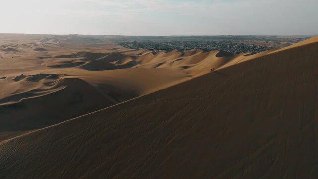 Aerial view of sand dunes in Huacachina desert, Peru, with a lush oasis surrounded by vast golden dunes under a clear sky. Scenic landscape showcasing natural patterns of wind-shaped sand formations.