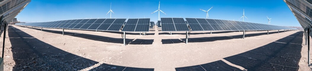 Panoramic view of hybrid plant with solar panels and wind turbines in Atacama Desert, Chile. Solar energy farm with photovoltaic panels and wind turbines under clear blue sky in arid desert landscape