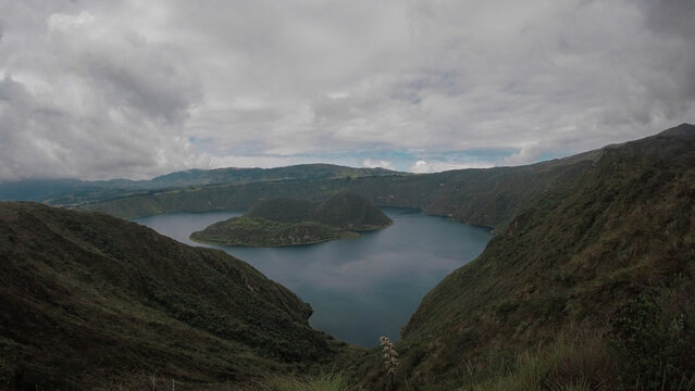 A stunning panoramic view of Laguna de Cuicocha, a deep blue crater lake within the Cotacachi Volcano. The image highlights its two distinct volcanic islands and the steep, lush caldera walls under a