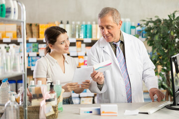Obraz premium Senior man pharmacist use computer at workplace, employee reads regulations and laws, makes report. Girl intern with prescription observes actions of professional, asks questions, practice work skills