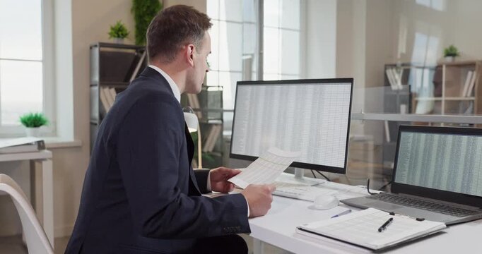 Man analyst in office working on computer. He reviews paperwork and data on monitor and laptop, checking spreadsheets and reports in a finance workspace. Concept of data driven analysis.