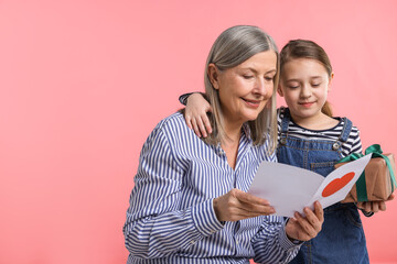 Little girl greeting her grandmother with holiday card and gift on pink background, space for text