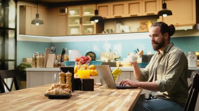 Portrait of male digital nomad using his laptop at the kitchen table, enjoying relaxed freelancer lifestyle with flexible work. Adult man telecommuting indoors, setup for productivity.