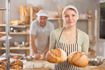 Bakery girl employee holds ready-made loafs of bread. He stands against background of bakerys desktop, flour, dough and improvised tools lie on work surface of countertop © JackF