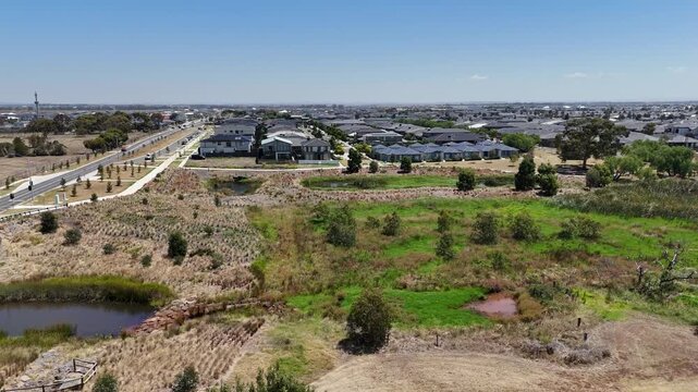 Aerial view of suburban housing estates in Tarneit, Melbourne, Australia, with stormwater wetlands and retention ponds integrated into urban neighborhood. A major road leads to expanding new estate