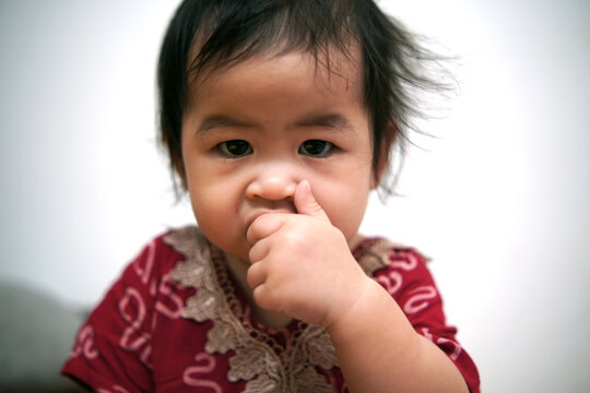 Adorable Asian toddler girl sucking her thumb with focused expression. Close up portrait of cute baby child with thumb sucking habit. Childhood psychology, self-soothing behavior