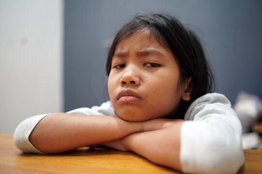 Moody Asian little girl sulking and leaning face on table, looking at camera with grumpy expression, unhappy child feeling bored or disappointed