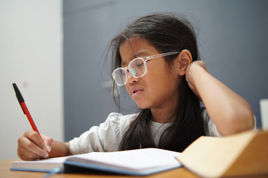 Stressed little Asian girl with glasses crying while studying, unhappy child having headache or difficulty with homework, academic pressure and learning disability