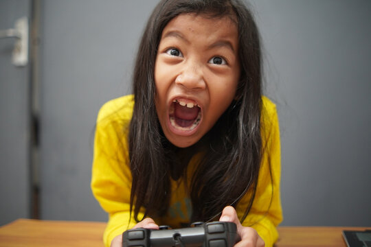 Frustrated little Asian girl grimacing while playing video games, upset child losing a game holding controller, gaming challenge and emotional reaction