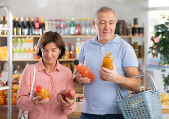 Fototapeta premium Couple of elderly man and elderly woman choosing natural juices in bottles in grocery store