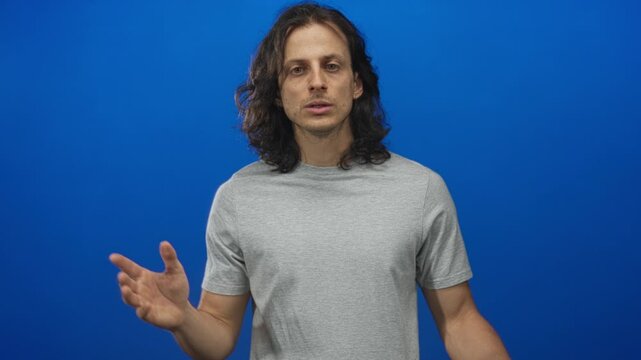 Man young hispanic with long hair pressing finger to lips in blue studio, facing camera with direct gaze; secrecy hush secretive.