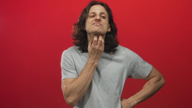 Young hispanic man scratches neck in red studio setting with visible hand and exposed neck gesture; irritation discomfort.