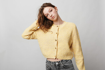 Young woman in yellow knitted cardigan and grey jeans poses with hand in hair against plain light...