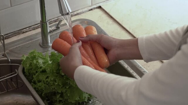 Hands washing carrots under tap, fresh orange carrots rinsed over stainless sink with green lettuce in colander, gentle scrubbing
