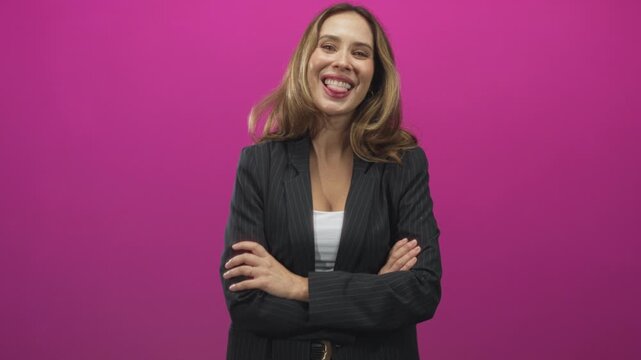 Woman with arms crossed smiling and sometimes sticking out tongue, wearing pinstripe blazer and white top in a pink studio; playful confidence approachable.