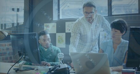 Leaning lead in checked shirt and pointing at open laptop while team reviewing monitors in office