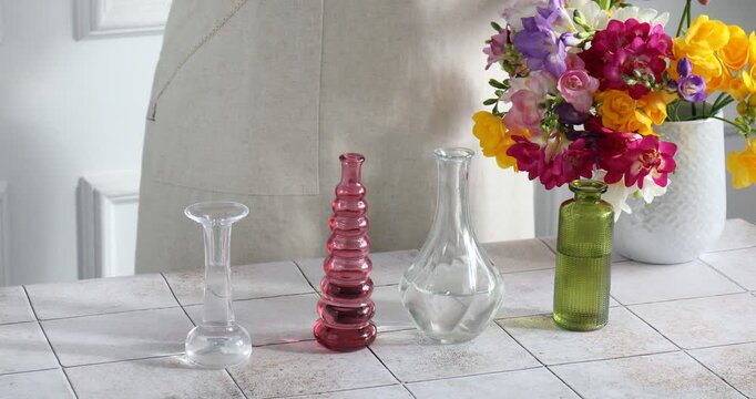 Woman putting beautiful freesia flowers into different vases at tiled table near white wall at home, closeup