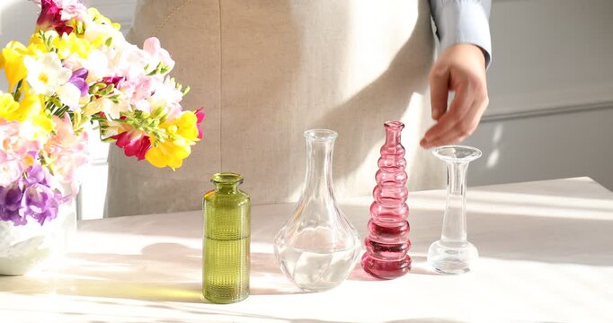 Woman putting beautiful freesia flowers into different vases at wooden table near white wall at home, closeup