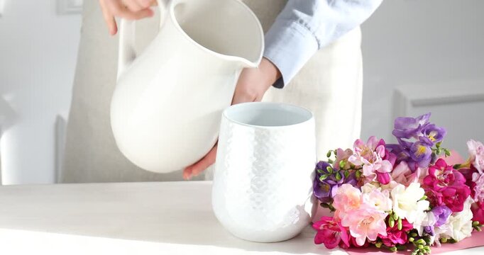 Woman pouring water from jug into vase and putting beautiful freesia flowers into it at wooden table near white wall at home, closeup