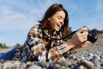 A woman photographer lies on pebbles at the beach, smiling as she frames with a camera. She wears a...