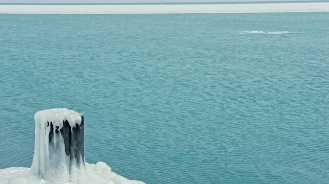A wooden post covered with ice is visible next to a frozen lake during winter. The water appears blue and calm with ice patches in the distance.