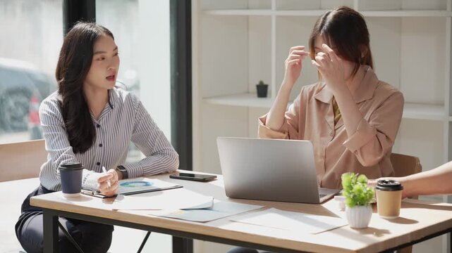 Two young Asian business professionals are sharing a laptop while sitting in a meeting, analyzing financial reports, using a calculator, and discussing data on charts.