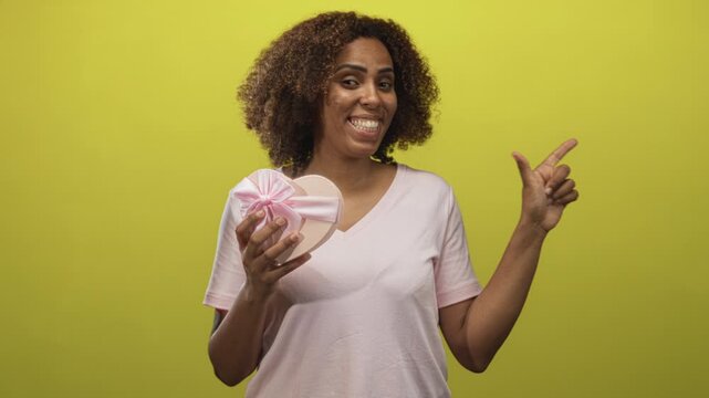 Young woman smiling while holding heart shaped gift box tied with pink ribbon and pointing finger in yellow studio; joy celebration love.