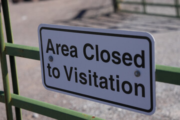 Fototapeta premium White rectangular “Area Closed to Visitation” sign is bolted to a green metal fence with a dry desert landscape and blue sky softly blurred in the background.