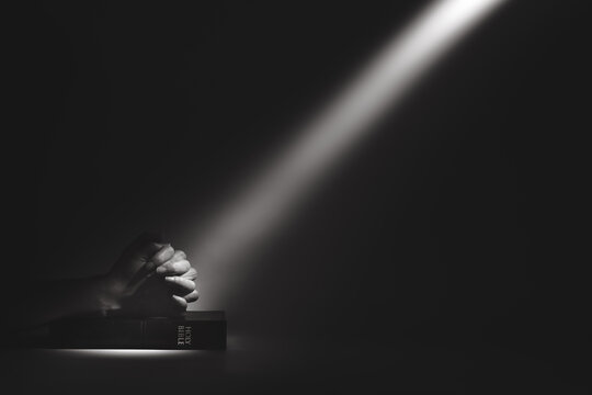 A strong beam of light illuminating a holy bible and the hands of a person praying, a background of faith and belief.
