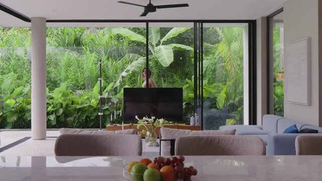 Modern living room interior with large glass windows showing lush green garden outside, cozy sofa, flat screen TV and decorative plants on table for home decor concept.