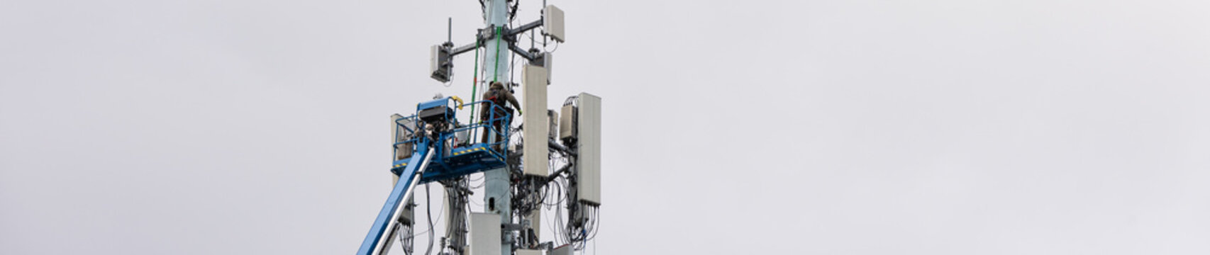 Boom truck lift backet with technician workmen raised up to antenna platform to perform maintenance, wireless mobile communication co-located cell sites on tall tower on cold winter day
