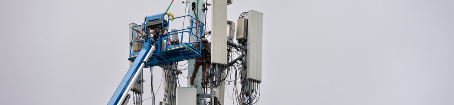 Boom truck lift backet with technician workmen raised up to antenna platform to perform maintenance, wireless mobile communication co-located cell sites on tall tower on cold winter day
