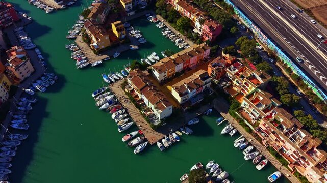 Boats are parked in a channel with green water surrounded by buildings. A busy road runs parallel to the marina. It is daytime with clear sunlight.