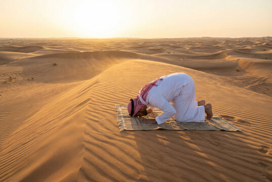 A man in traditional attire prostrating on a prayer mat in the desert at sunset, performing a religious prayer.