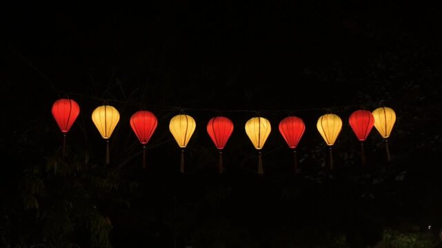 Glowing Red Yellow Lanterns Strung In Hoi An Night Riverside, Warm Illumination Over Water, Paper Lanterns Swinging Gently, Dark Foliage Silhouette, Calm Festive Atmosphere, Horizontal Row Of Lights