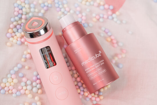 Bar, Montenegro: February 6, 2026: Pink handheld facial device with orange Booster Mode LED ring beside a rose gold medicube collagen serum bottle on soft pastel fabric